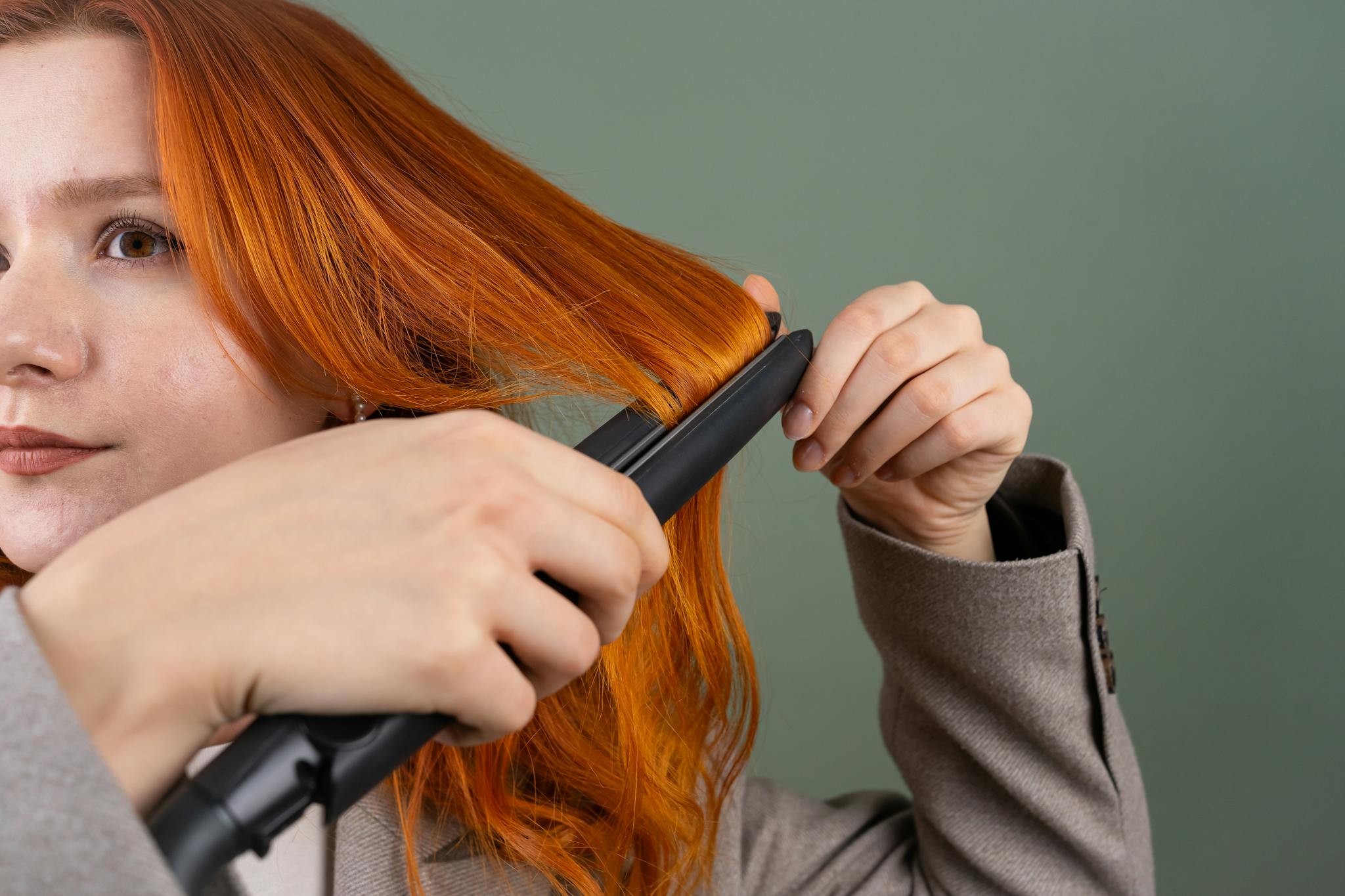 Home Close-up of a woman styling her red hair using a hair straightener against a green background.