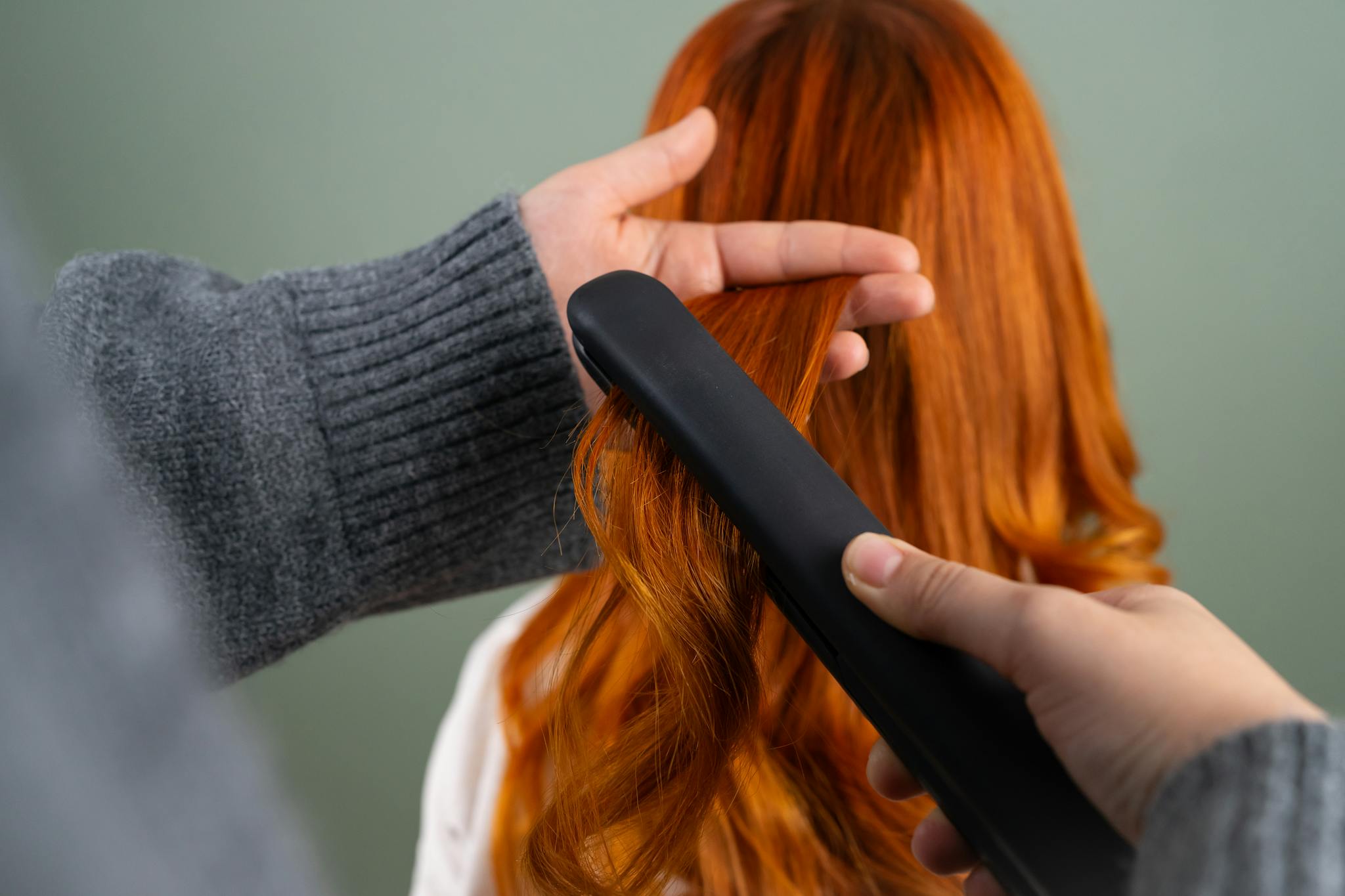 Home Hands styling long red hair using a flat iron indoors. Haircare and beauty concept.